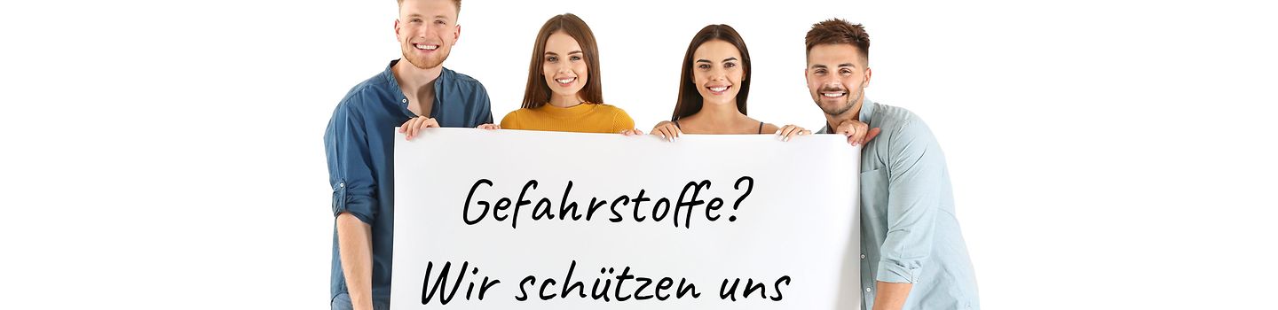 Vier junge Leute halten vor sich ein großes weißes Schild. Auf dem Schild steht "Gefahrstoffe? Wir schützen uns" in schwarzen Buchstaben